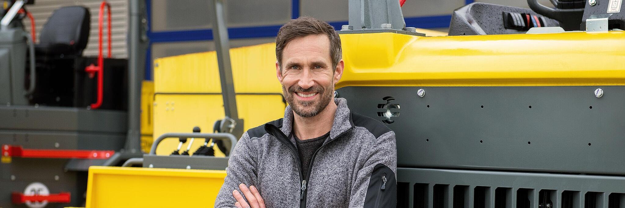 Un empleado sonriente de Wacker Neuson se apoya en una máquina para la construcción de Wacker Neuson con los brazos cruzados.