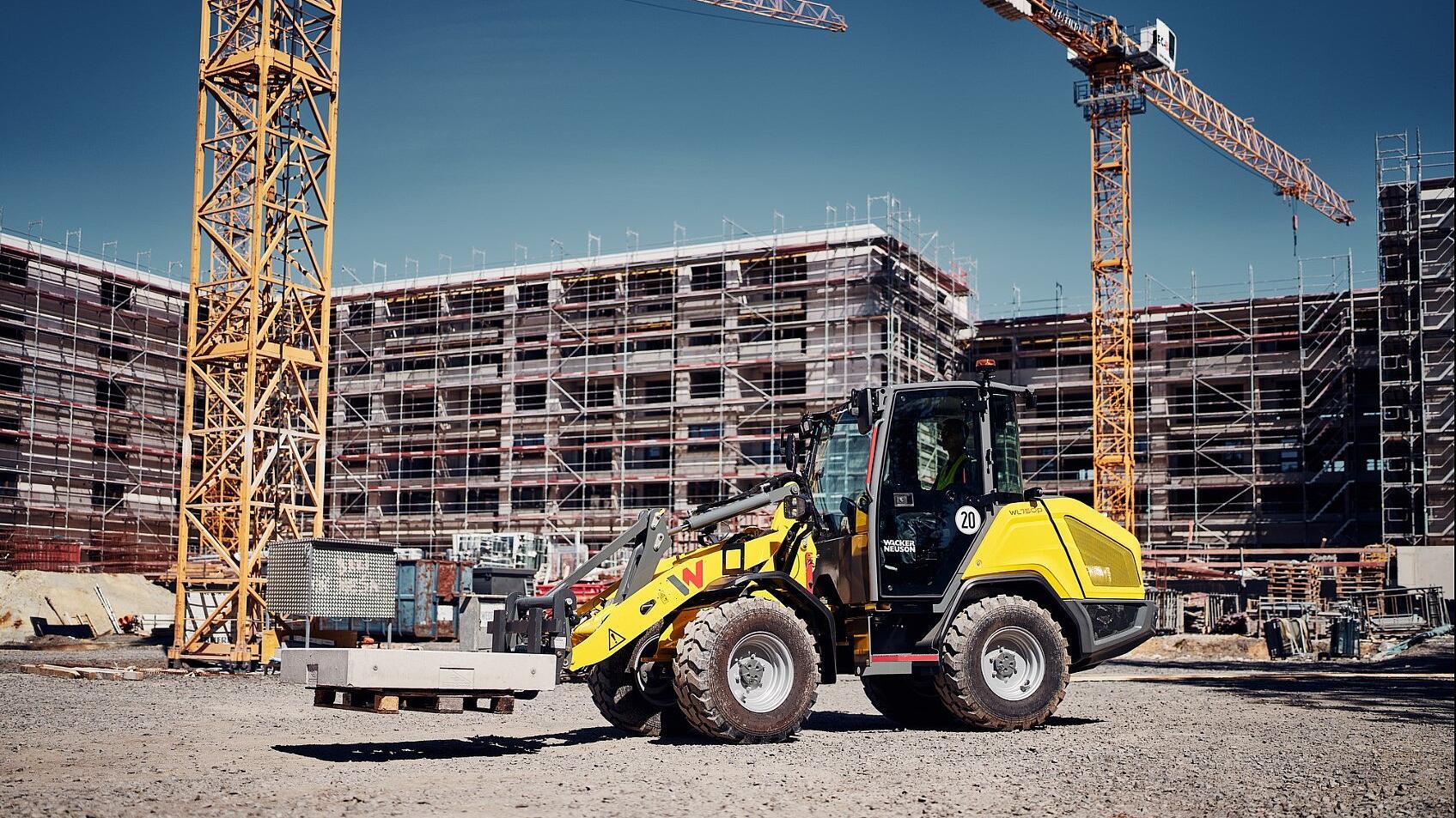Wacker Neuson WL750p wheel loader transporting concrete elements across an active construction site with scaffolding and tower cranes in the background.