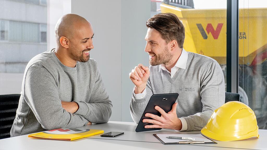 Funcionário da Wacker Neuson sentado à mesa de trabalho mostrando algo a um cliente no tablet.